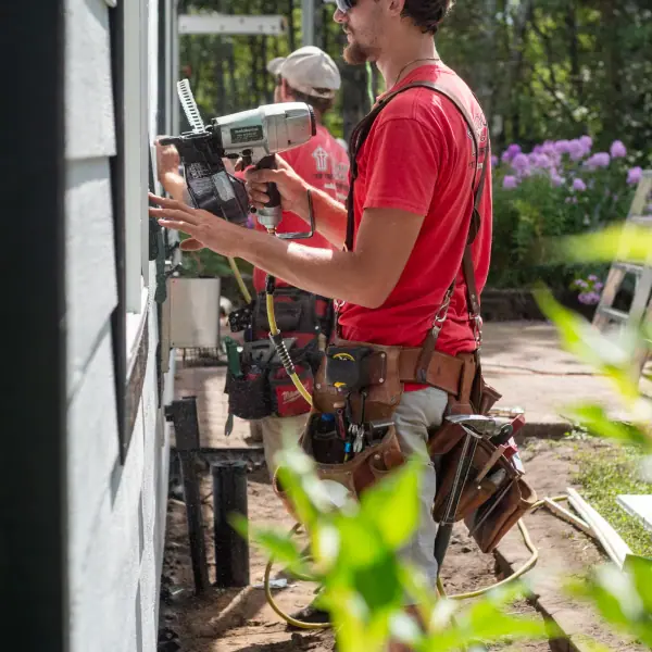 A worker holds a pneumatic coil nailer against a white lap siding wall during an exterior installation.