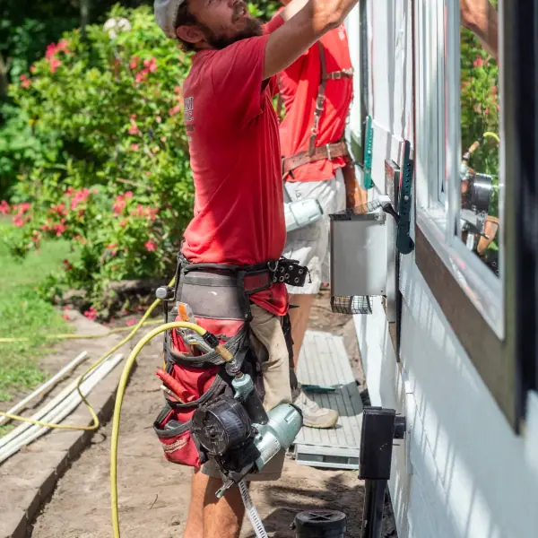 Two siding installers in red shirts lift a white lap siding board into place above a window on a sunny day.