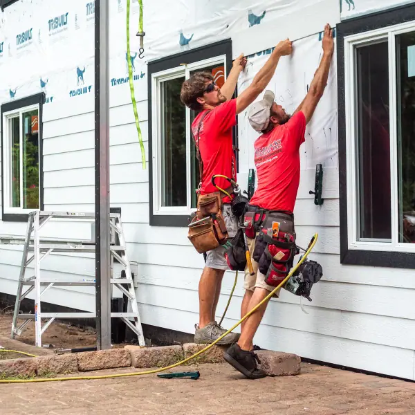 Two installers lift a siding panel into place over housewrap beside black-trimmed windows.