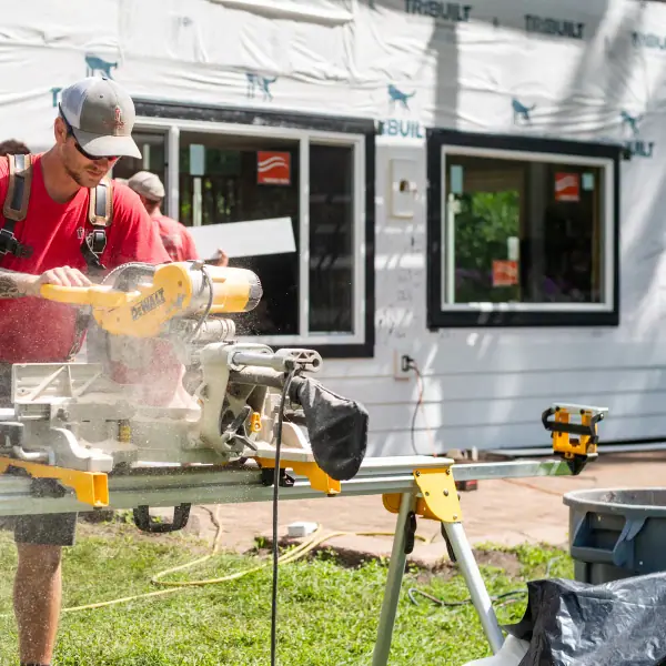 A crew member cuts trim on a miter saw, sawdust flying, in front of a partially sided home.