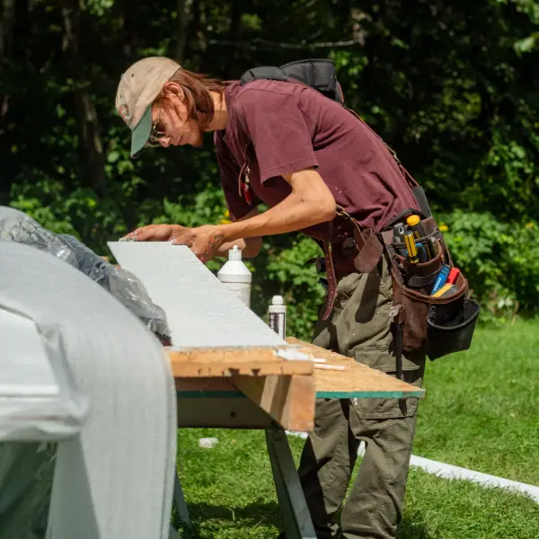Crew member working on a white trim board at an outdoor bench with a tool belt and sealant nearby.